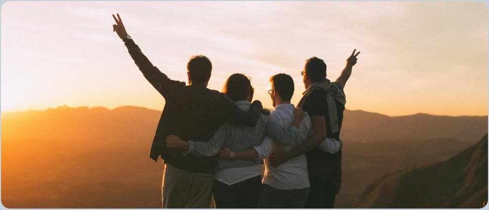 Four friends with their arms around each other, backs to the camera, raising their hands as they watch the sun set over a mountain valley